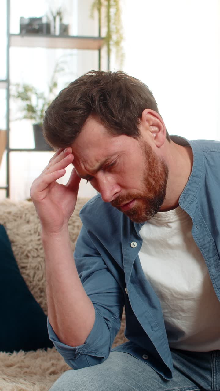 Young man sitting on the sofa at home holding his head and massaging temples due to intense headache