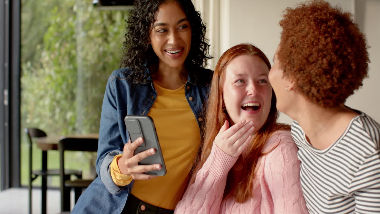 Showing smartphone, diverse female friends laughing and enjoying time together indoors