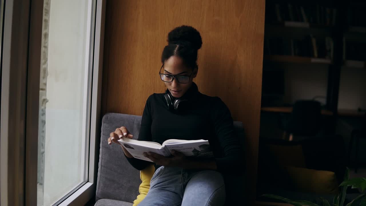 The young attractive afro-american woman is reading the book on the wide window-sill. Close-up portrait of a stylish girl in eyeglasses and headphones reading or studying