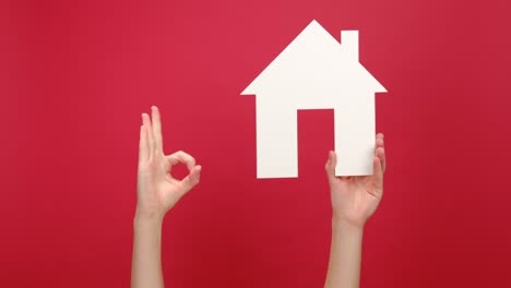 female hands holding white house model and makes okay gesture, real estate home approval, isolated over red color background wall with copy space for advertisement. concept social service about home