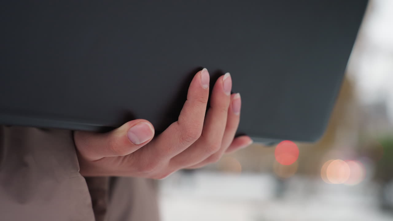 Close-up view of light-skinned hand gently holding black laptop against soft blurred background with glowing red bokeh lights, suggesting focus and outdoor winter setting in subtle natural lighting