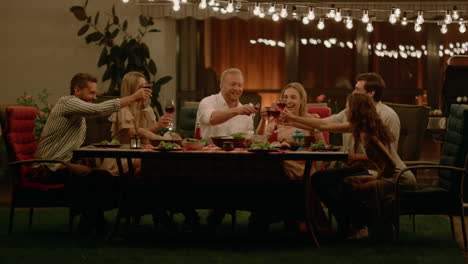 Friends-sitting-at-dining-table-on-backyard-of-house.-Positive-man-making-toast