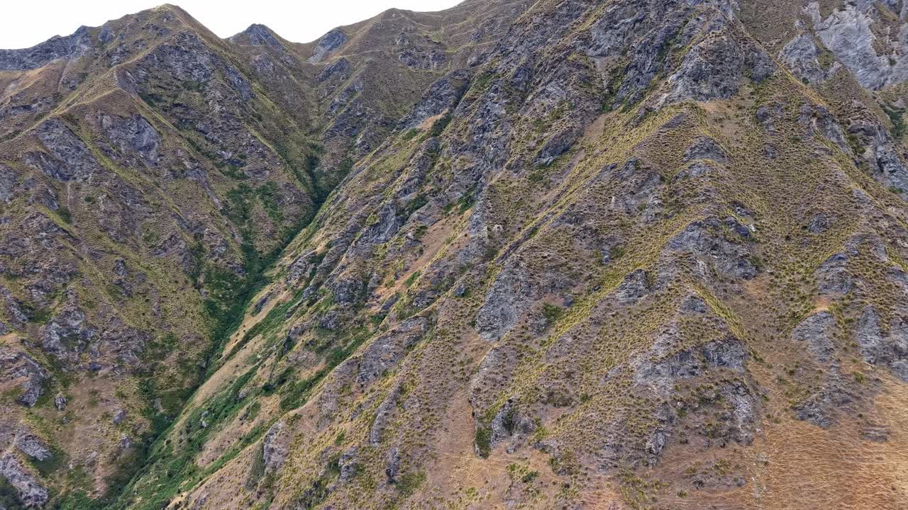 Drone footage approaching the rugged mountains of Roys Peak hiking track in Wanaka, New Zealand, showing steep slopes and rocky textures