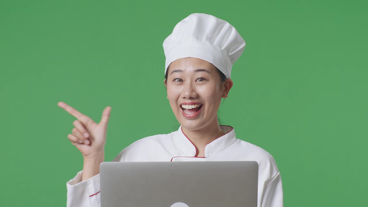 Close Up Of Asian Woman Chef Using A Laptop And Pointing To Side While Standing In The Green Screen Background Studio