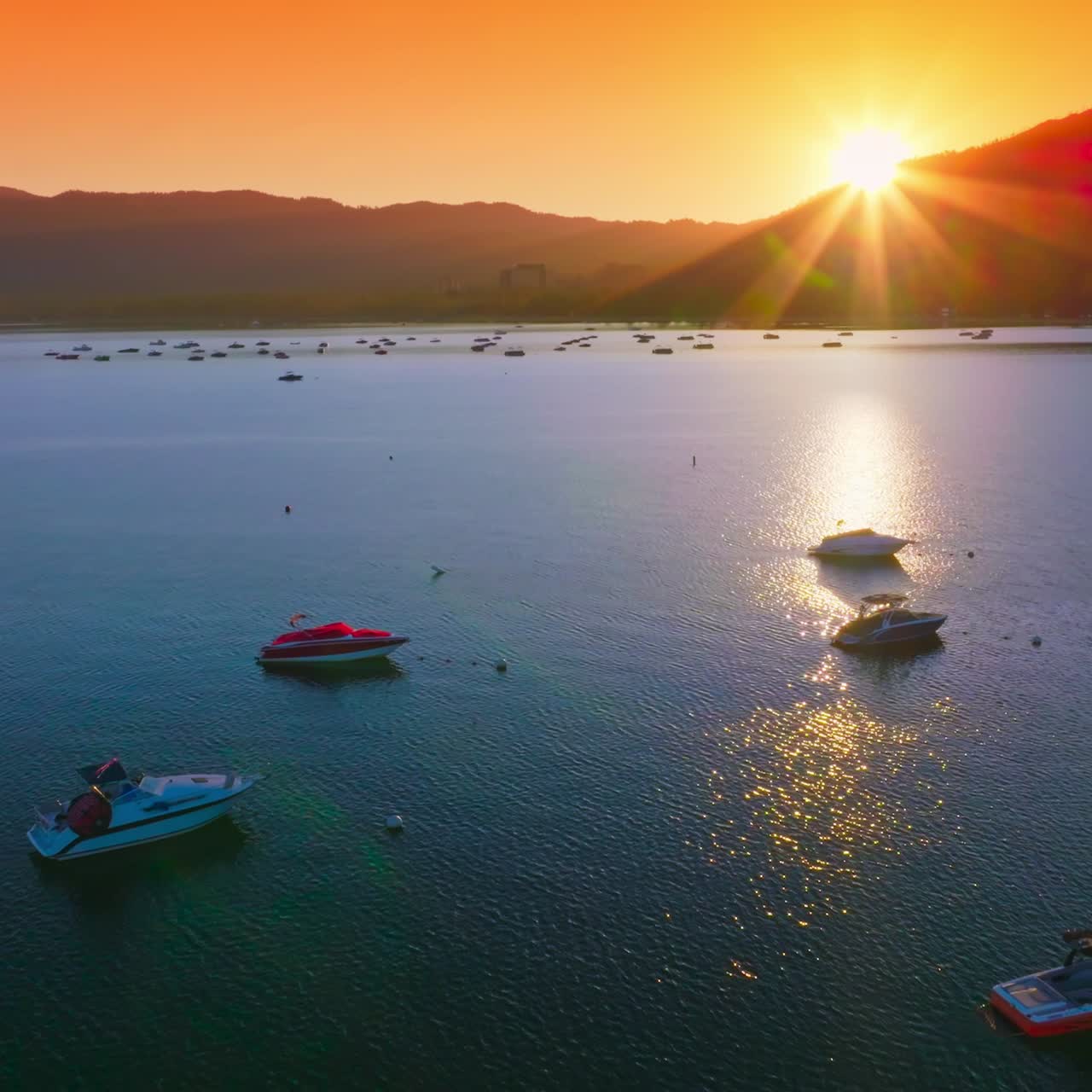 Sun sending last rays from behind the mountains. Multiple yachts and boats standing on anchor on the lake. Orange sky at backdrop