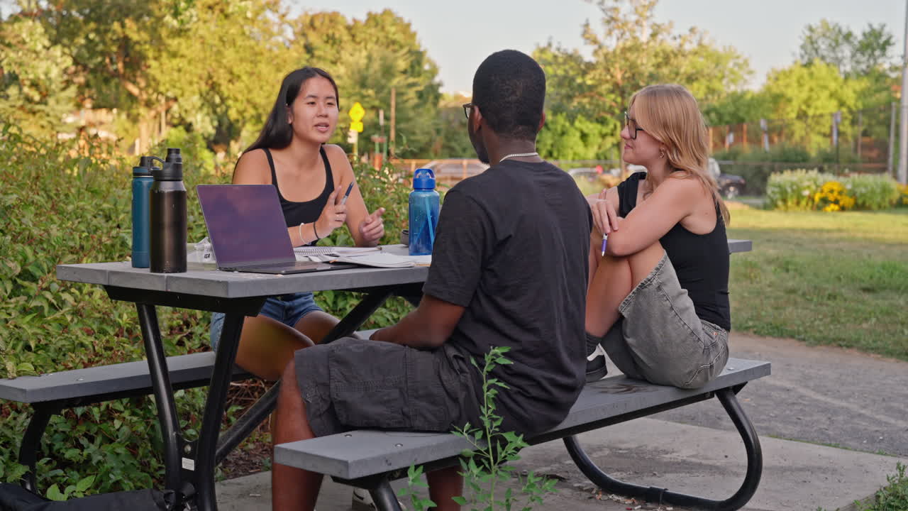 Happy multicultural group of students with many interactions, studying outdoors at park table with laptop and notebooks, symbol of diversity, teamwork and education