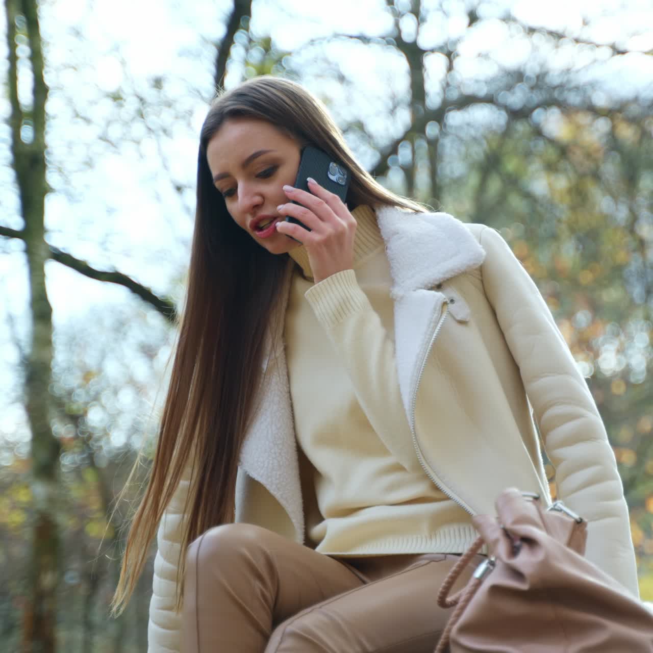 Affascinante donna bruna che parla al telefono nel parco autunnale. Donna seduta nella natura che chatta con qualcuno. Angolazione dal basso