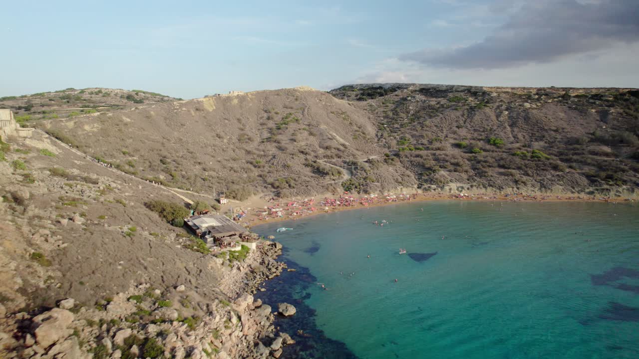 The drone flies close to the rocky cliffs of Golden Bay, passes over a tower under restoration, and reveals the sunny beach and anchored boats below