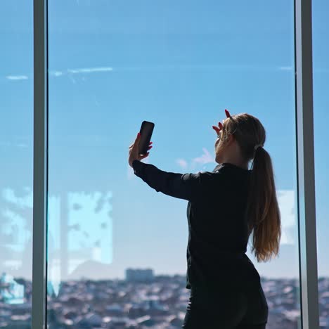 lovely business woman in black taking photo of herself. beautiful young woman doing selfie on smartphone while standing at office window