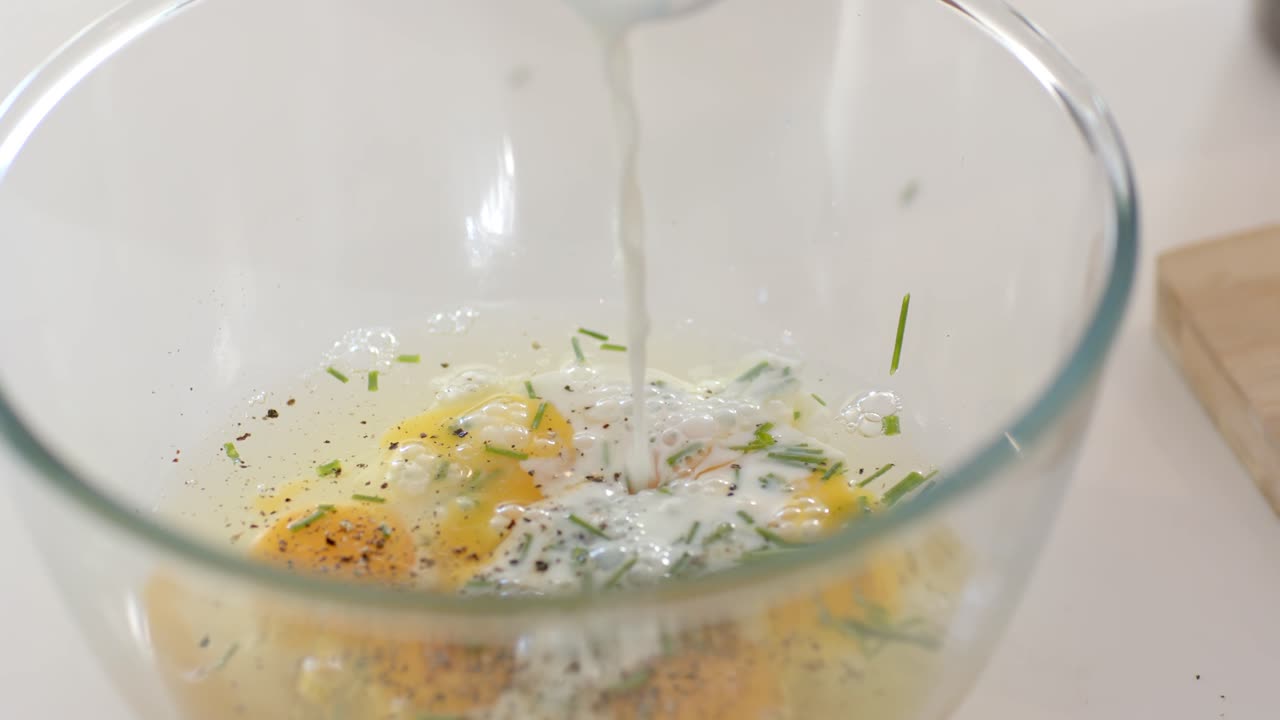 Glass mixing bowl with three eggs, chives and pepper, milk pouring from spoon at kitchen counter