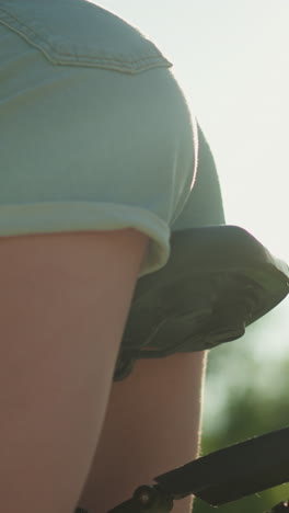 vista lateral de una mujer ciclista en pantalones cortos de vaqueros azules ajustándose en el asiento de la bicicleta, moviéndose ligeramente para la comodidad, escena iluminada por el sol con vegetación y fondo suave borroso