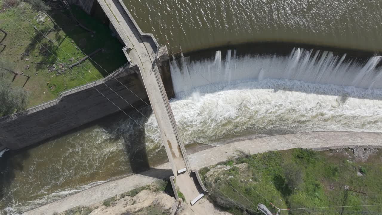low motion top-down drone footage of a dam spillway with a lateral flight view. A bridge appears as water cascades down powerfully through a wide channel. The scene is truly spectacular.