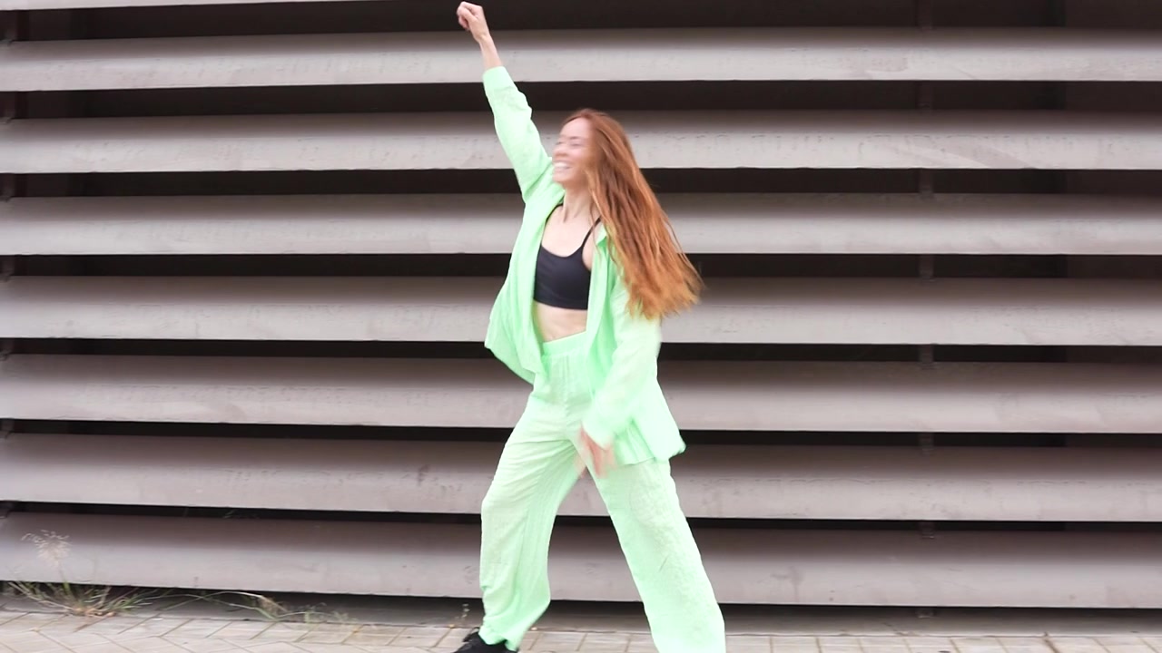 Mujer con atuendo verde neón bailando enérgicamente frente a un fondo industrial.