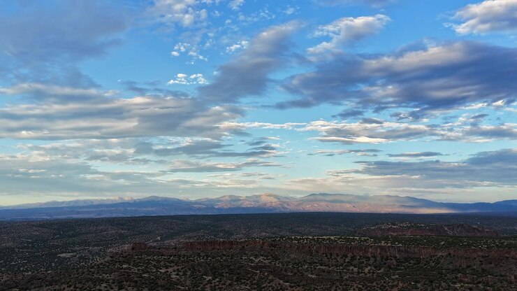 Timelapse 4K con dron sobre el vasto desierto alto del Condado de Santa Fe, mirando hacia la distante cordillera de Sangre de Cristo y el Monte Baldy. Suave luz de atardecer y nubes dispersas flotan sobre el terreno accidentado