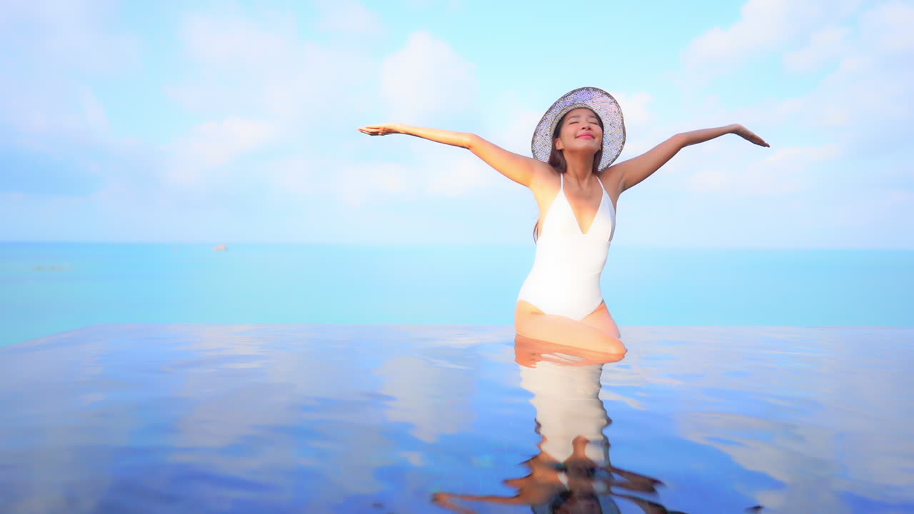 수영장 가장자리에 앉아 있는 젊은 매력적인 여성이 기쁨에 팔을 들어 올립니다