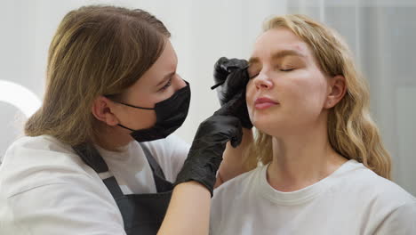 beautician wearing gloves and face mask attentively picks client eyebrow with precision as client keeps eyes closed. scene set against soft white curtain background, highlighting calm interaction