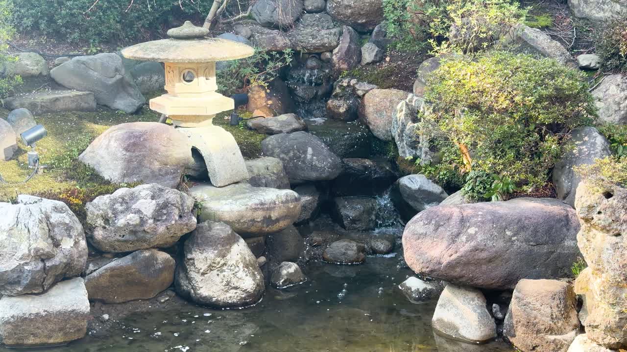 Ampia inquadratura fissa di una lanterna di pietra e un laghetto roccioso in un giardino di Kanazawa.