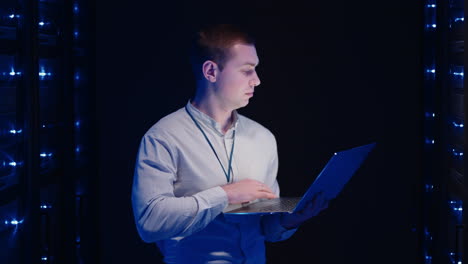 young man holds device in hand and looks at screen inspects equipment or hardware rack. male programmer working with laptop and supporting service while standing in data center spbas.