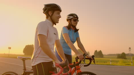 a man and a woman in helmets with bicycles stand and talk at sunset. rest after a bike ride on the highway. track bikes. couple sports