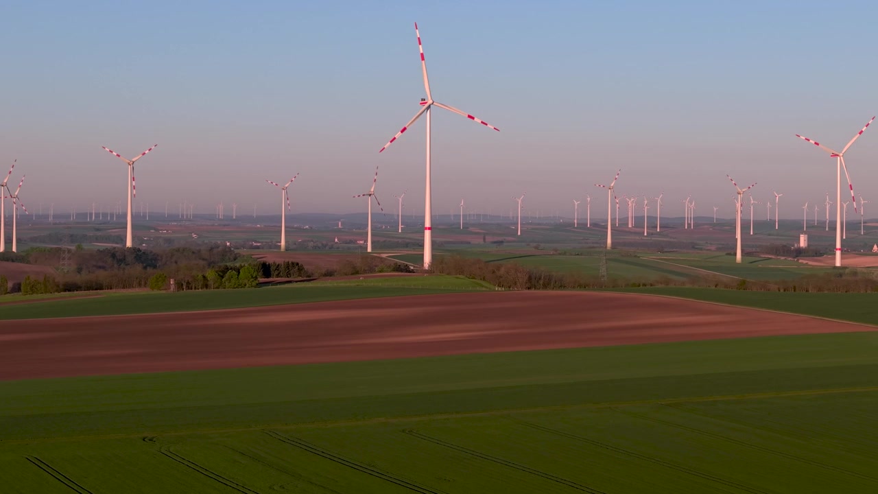 Luchtfoto van een groot windmolenpark bij zonsopgang in het gouden uur licht. Meerdere witte windmolens met rode markeringen verspreid over een agrarisch landschap. Installatie voor hernieuwbare energie op het platteland.