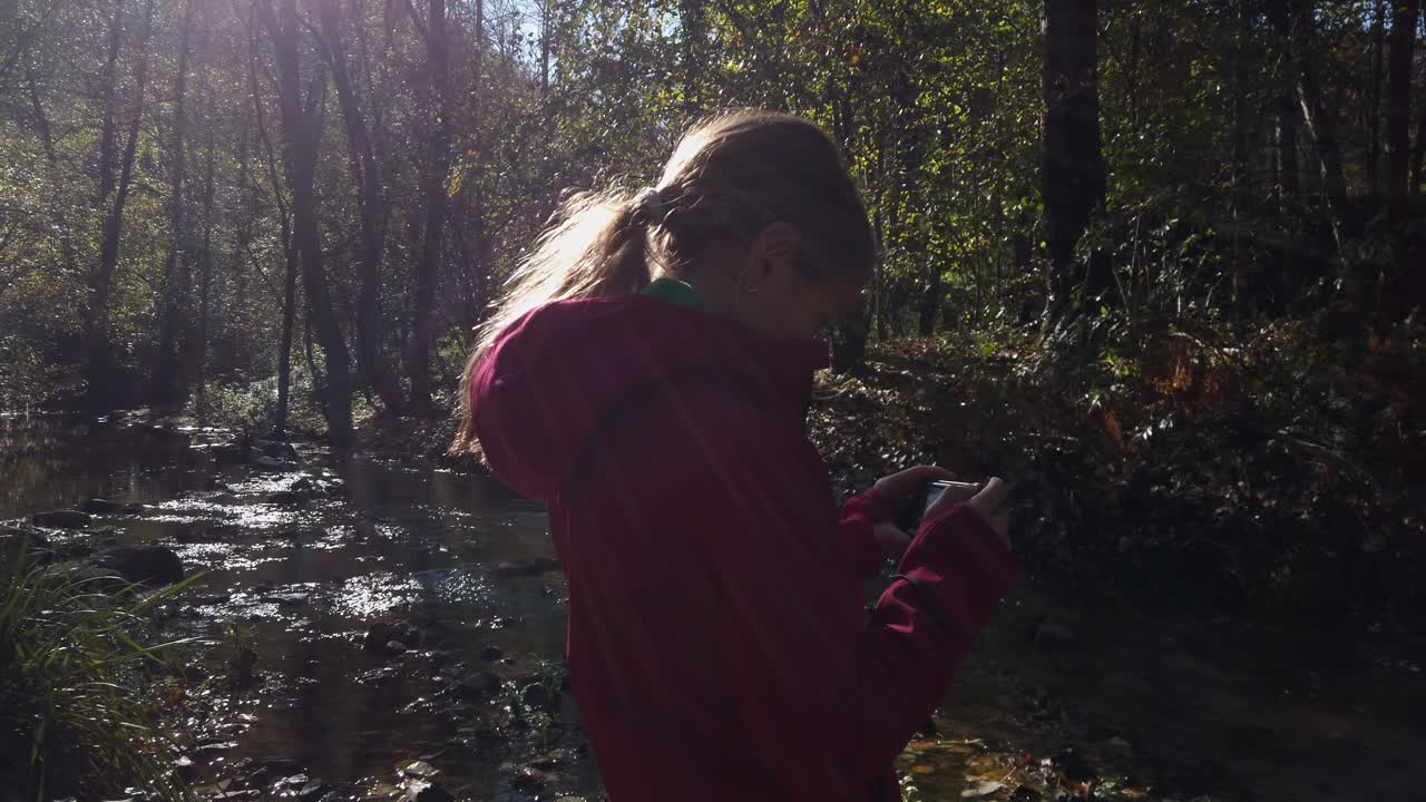 Young girl, wearing a red jacket, is engrossed in taking pictures with her digital cam by a gently flowing river in a serene forest setting, sunlight filters through the trees