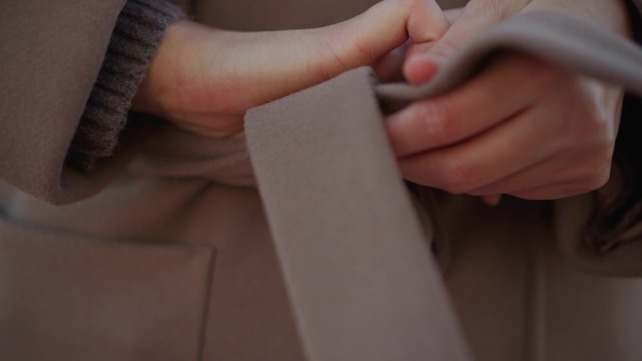 Primo piano di donna che sistema la cintura del suo cappotto marrone, concentrandosi sulle mani e sulla texture del tessuto. Il soggetto manipola il materiale morbido con movimenti intenzionali delle mani in un ambiente esterno con sfondo sfocato.