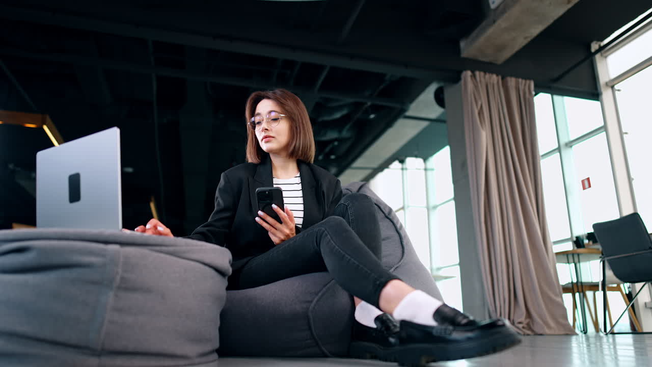 Freelancing lady uses laptop and her smartphone. Woman sits in bean bag chair using her computer. Low angle view.