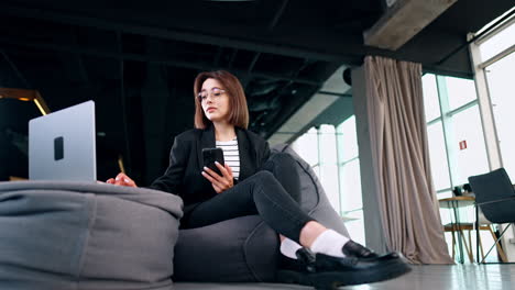 freelancing lady uses laptop and her smartphone. woman sits in bean bag chair using her computer. low angle view.
