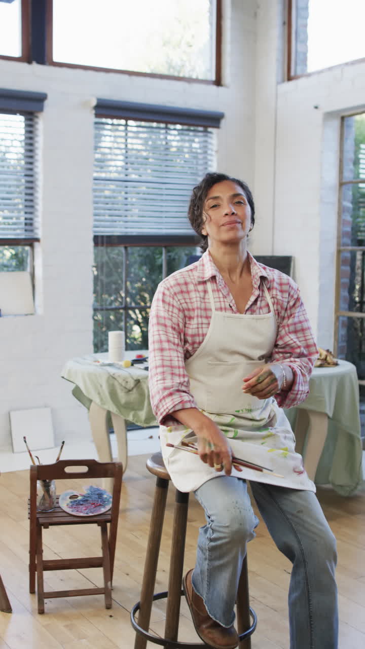 Vertical video of portrait of happy biracial woman in apron holding brushes in studio, slow motion