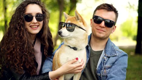 proud owners are putting sunglasses on adorable shiba inu puppy, laughing and chatting relaxing in city park in summer. people, animals, nature and fun concept.