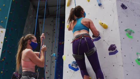 two caucasian women wearing face masks using ropes to climb wall at indoor climbing wall