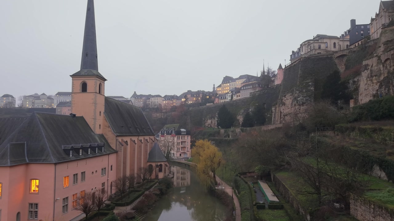 Drone flight over the Alzette River in Luxembourg, capturing Neumunster Abbey on one side and the city rising over the hill on the other, revealing a scenic historic urban landscape.