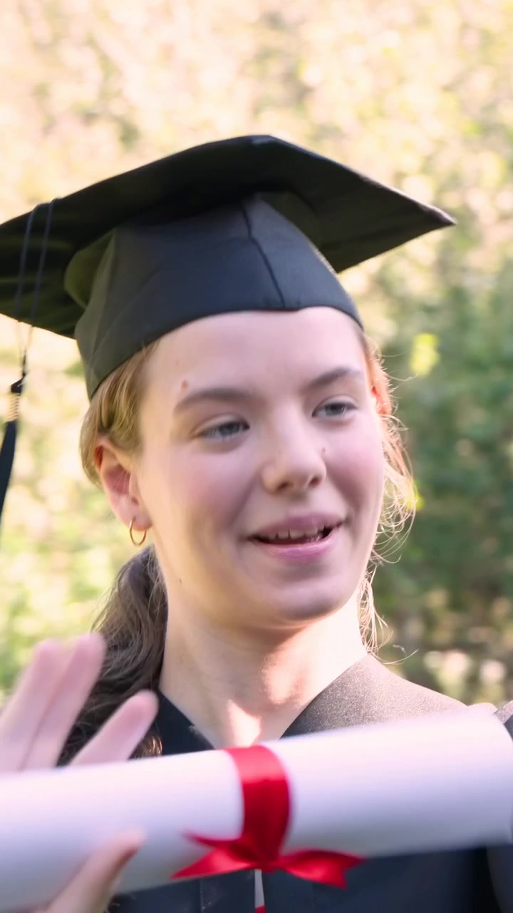 Vertikal video: Smilende dimittend vender sig og vifter med diplom efter et signal uden for billedet i en have, iført kasket.