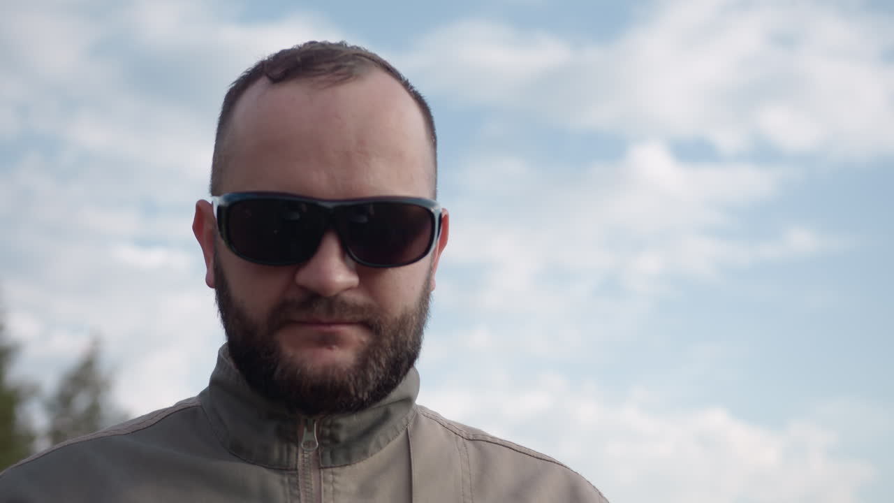 close up of man wearing dark sunglasses gazing into sky above blurred trees, beard outline defined against bright clouds, pensive expression captured in soft light, nature backdrop creating calm mood