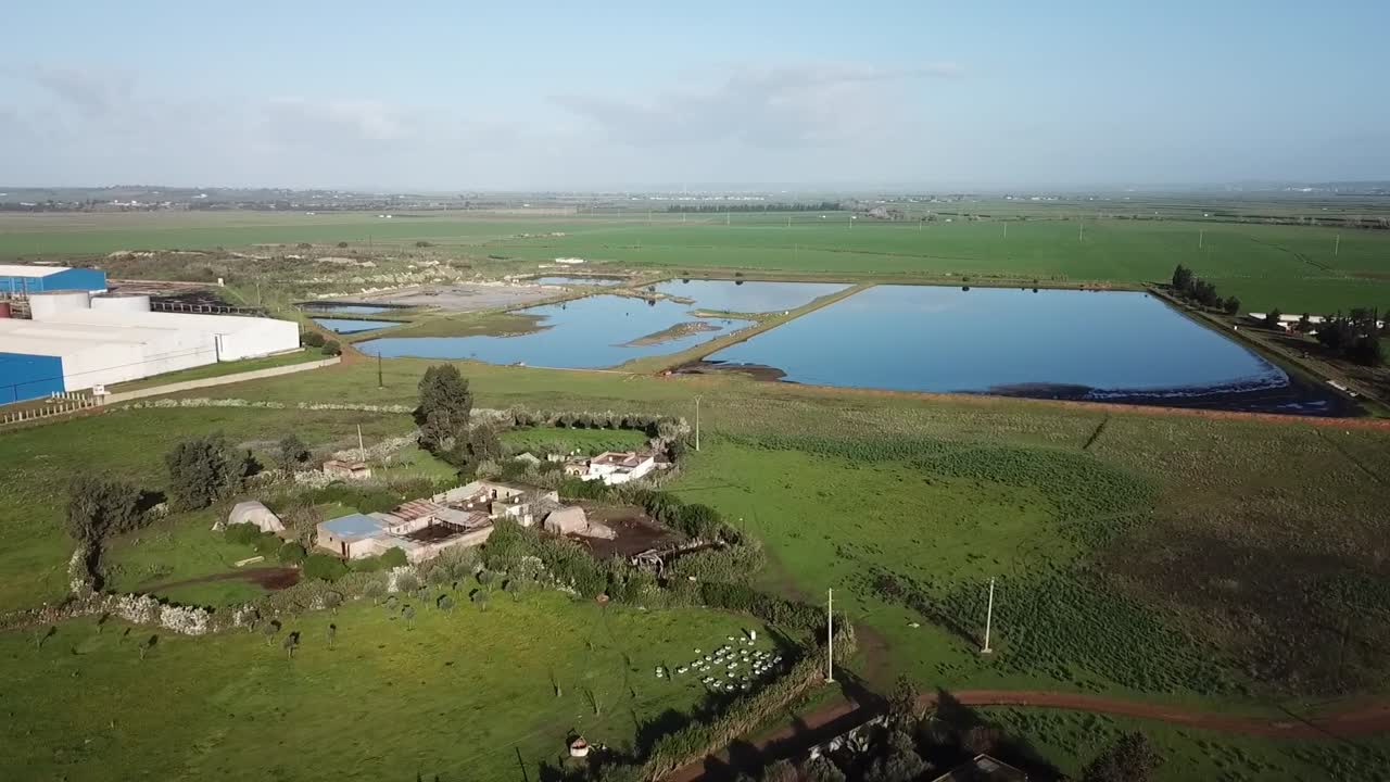 Prise de vue par drone d'un bassin de stockage d'eau stratégique pour l'irrigation. Montre le village rural environnant, une usine agricole industrielle moderne et de vastes champs fertiles dans le Nord du Maroc.
