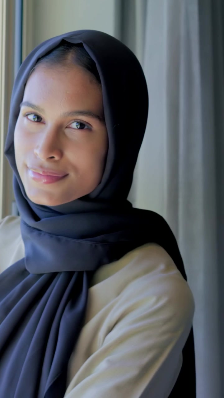 Vertikal video: Kvinde med blikket rettet, i sort hijab og beige top, bevæger sig ved vinduet efter en pause og viser ur