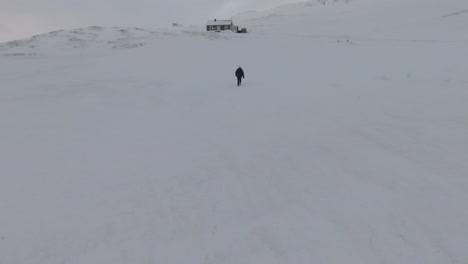 Dolly-forward,-woman-walking-towards-snowed-cabin,-white-mountains-background