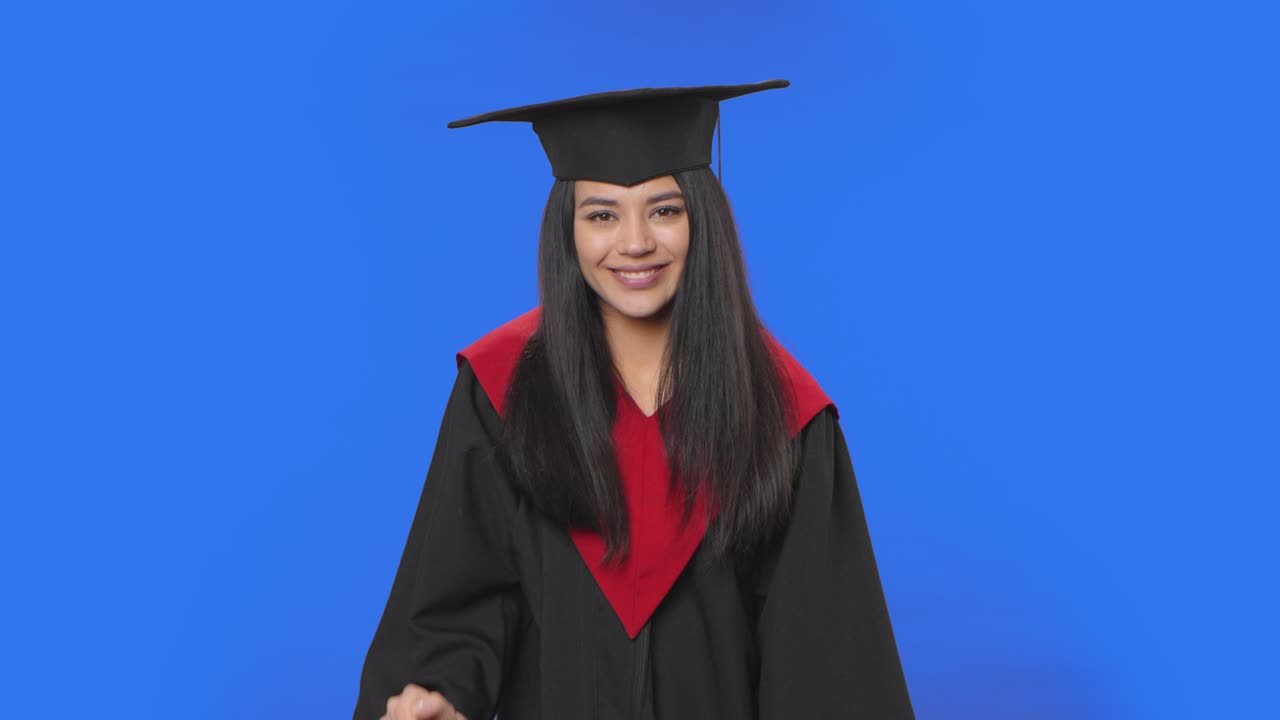 Portrait of female student in cap and gown graduation costume smiling and blowing kiss. Young brunette woman posing in studio with blue screen background. Close up. Slow motion ready 59.94fps