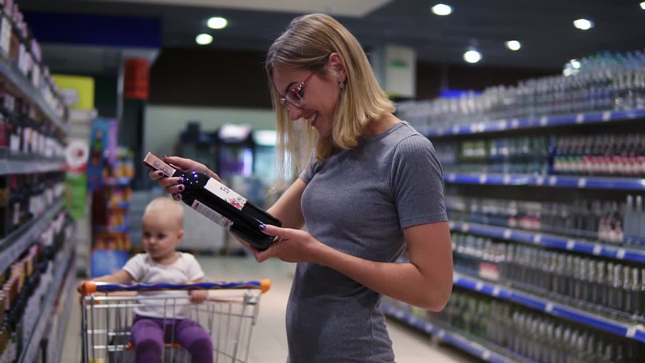 Attractive woman in glasses is choosing a bottle of wine in beverages department in the supermarket, while her little baby is