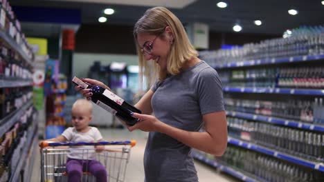 Attractive-woman-in-glasses-is-choosing-a-bottle-of-wine-in-beverages-department-in-the-supermarket,-while-her-little-baby-is