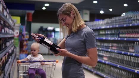 attractive woman in glasses is choosing a bottle of wine in beverages department in the supermarket, while her little baby is