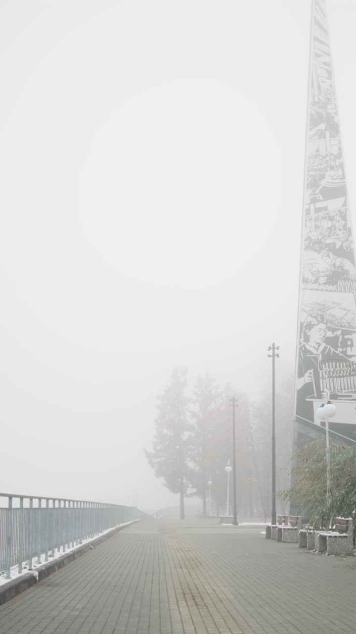 Winter cityscape flooded with dense fog, snowfall dusting park benches and frosted trees lining quiet walkway by iron railings and light poles, distant mast looming blurred
