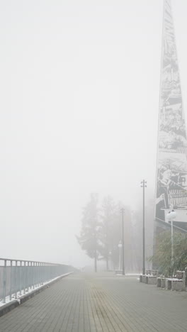winter cityscape flooded with dense fog, snowfall dusting park benches and frosted trees lining quiet walkway by iron railings and light poles, distant mast looming blurred