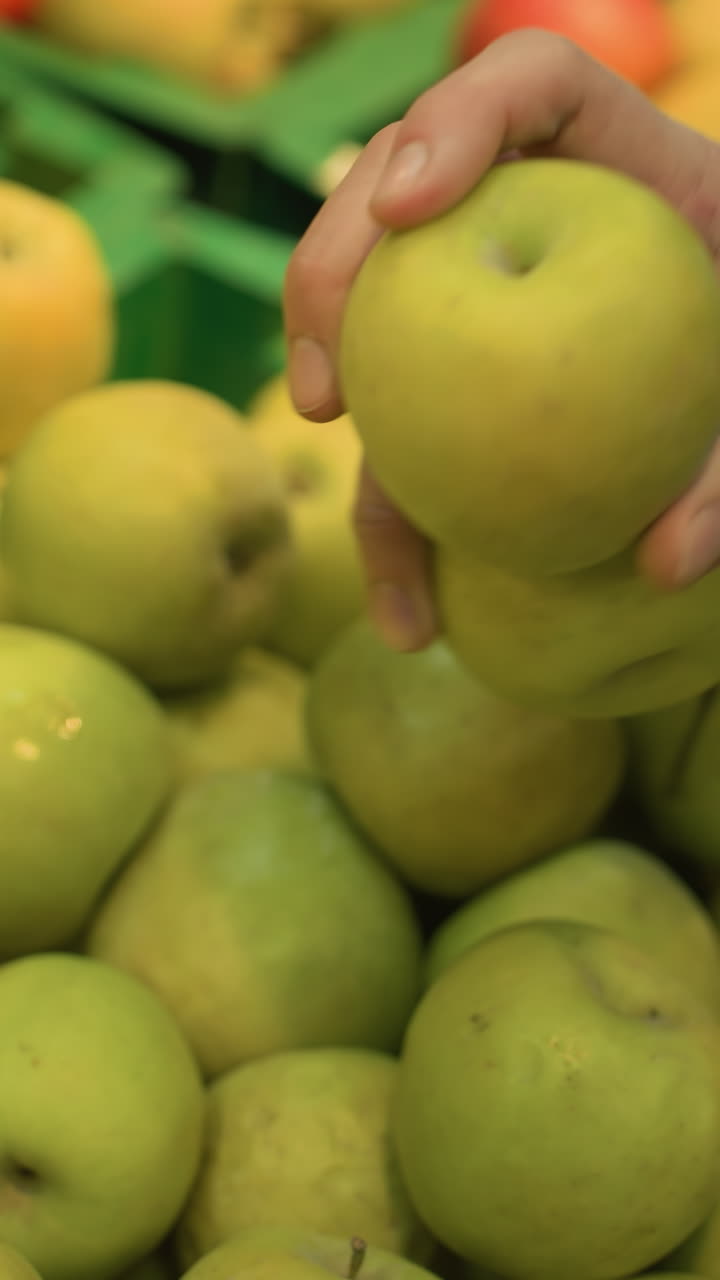 view of a hand wearing bead bracelets picking up two apples from a display in a grocery store. Shot with a handheld camera, capturing the detailed action in the produce section