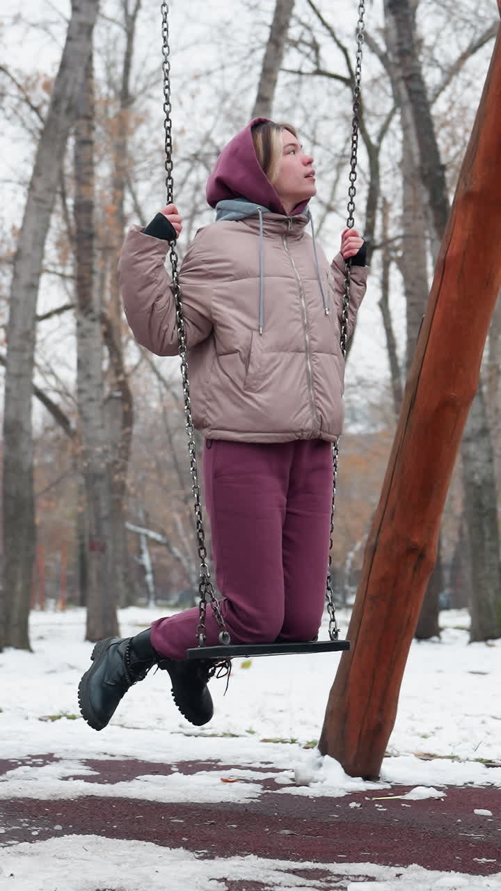 Giovane donna in abbigliamento invernale appoggia con cautela il ginocchio su un'altalena di ferro mentre si tiene alle catene per l'equilibrio, terreno coperto di neve, alberi spogli ed edifici residenziali sullo sfondo