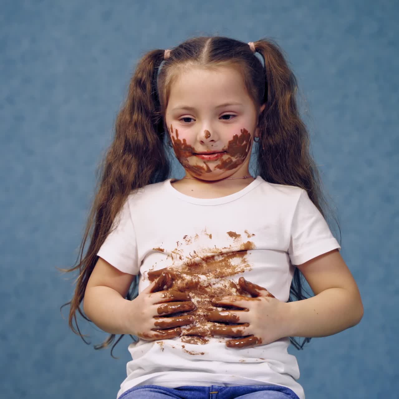 Portrait of dirty little girl after eating chocolate. Funny child with two ponytails have smeared face and white t-shirt in chocolate.