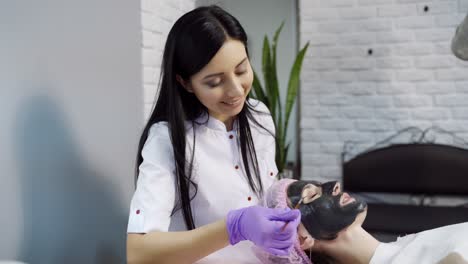 a cosmetologist with long hair stands behind the client's head and evenly distributes the cleansing mask on the face of the woman on the background of the office interior.
