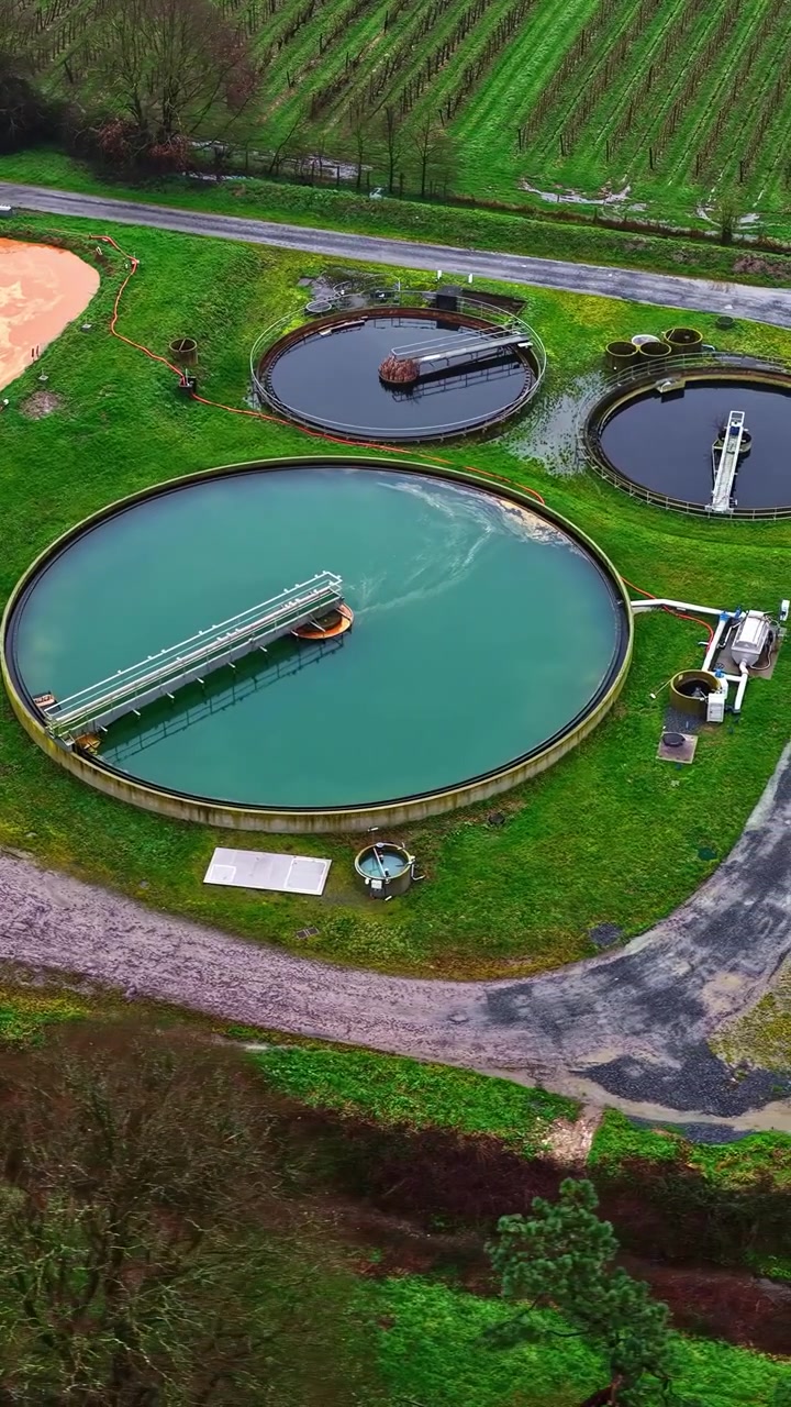 Colorful wastewater treatment basins beside farmland overview from above on cloudy day, aerial vertical of spinning pools