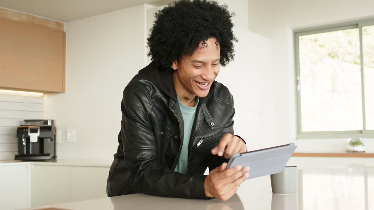 Uomo afroamericano che reagisce a un avviso del tablet al bancone della cucina, toccando lo schermo e ridendo accanto a una tazza