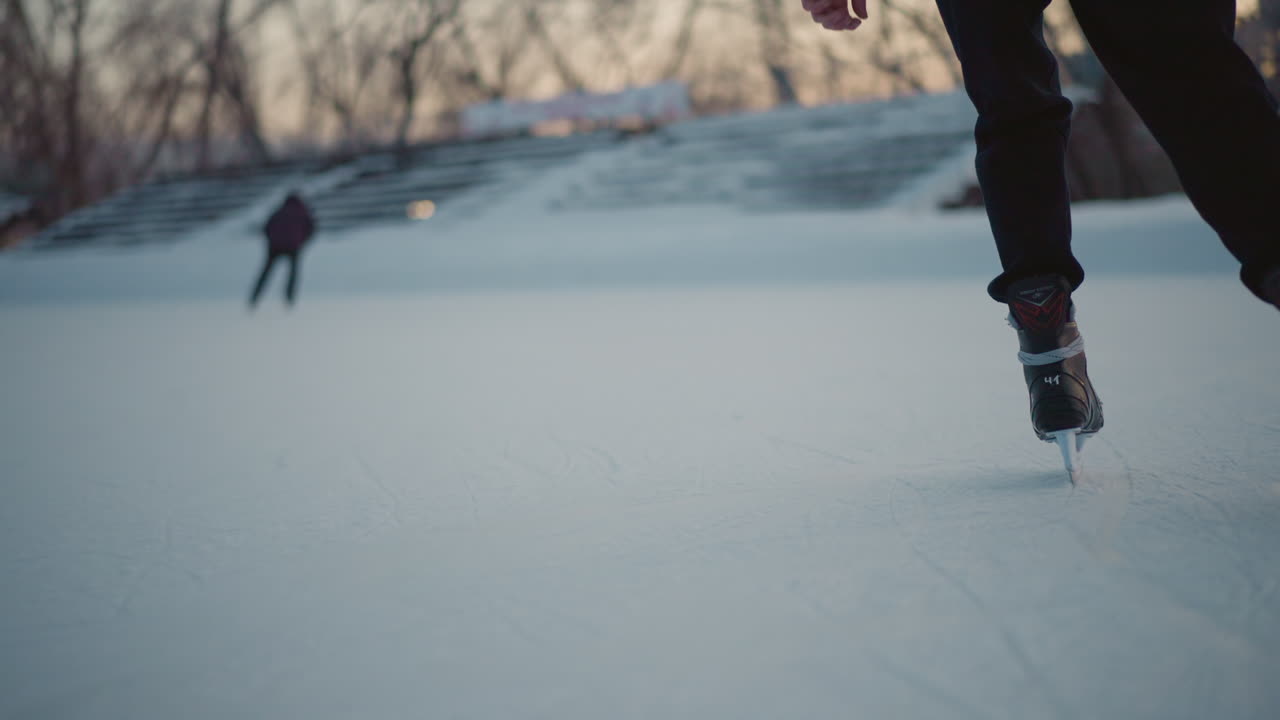 rear view close up of skater gliding on frozen lake wearing black ice skates laced with gray straps illuminated by soft evening light with blurred background of snowy stadium and trees in distance